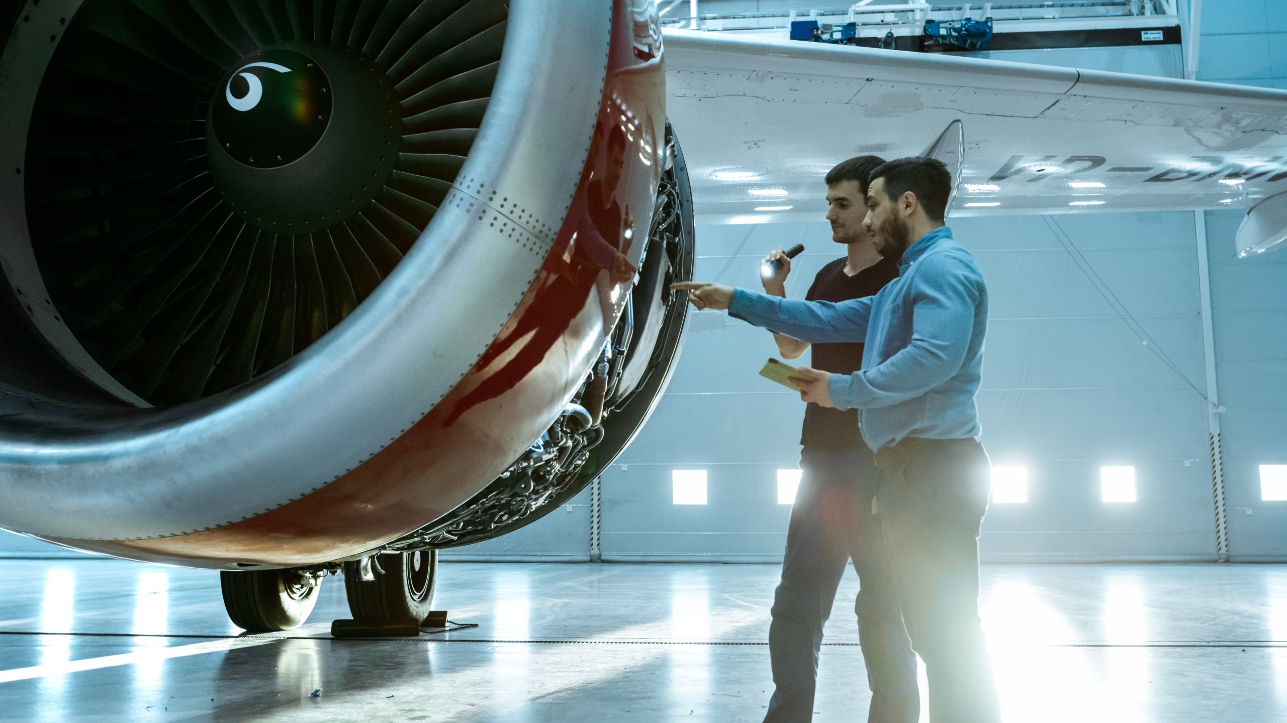Two individuals examining a large aircraft engine in a brightly lit hangar.