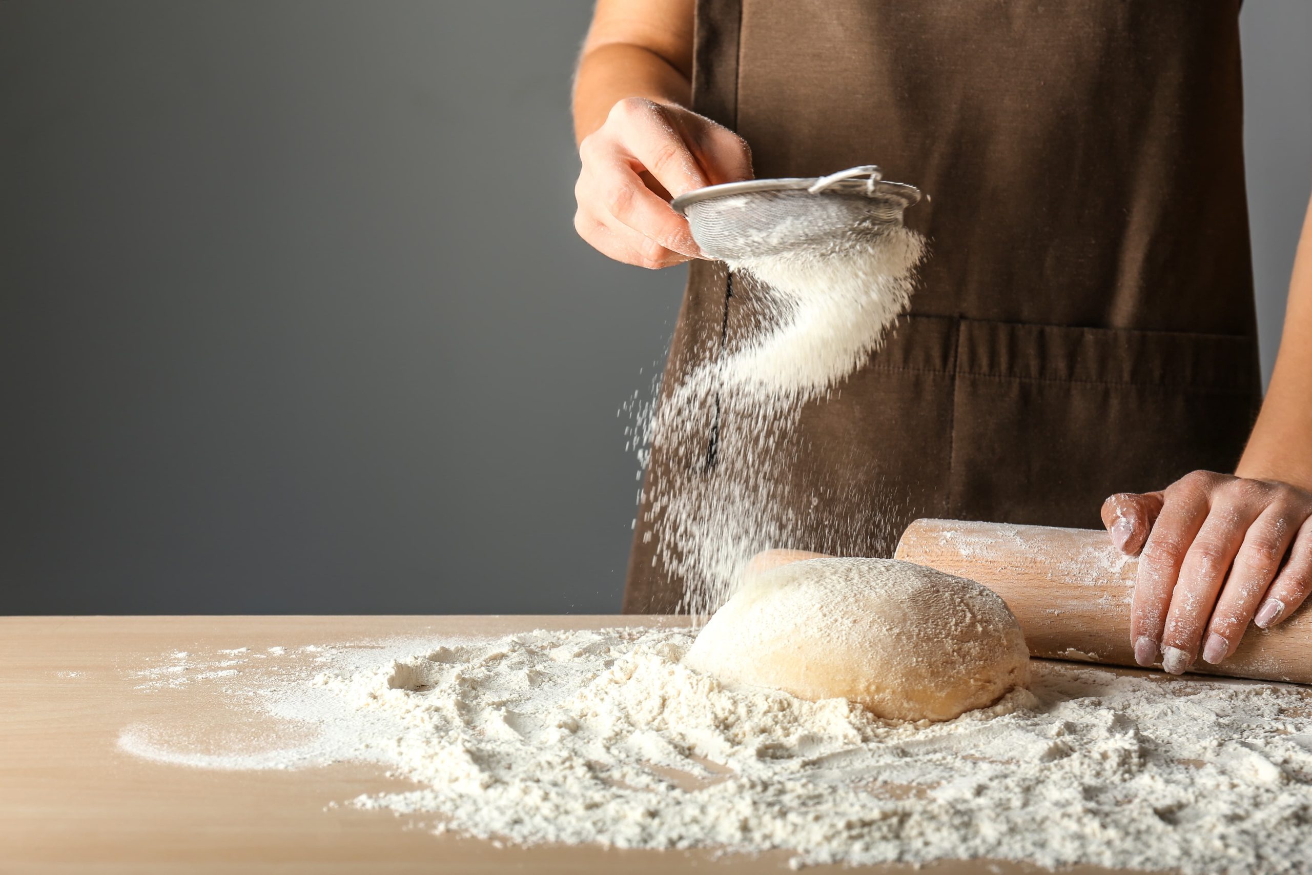 A person sifting flour over dough on a wooden table with a rolling pin nearby.