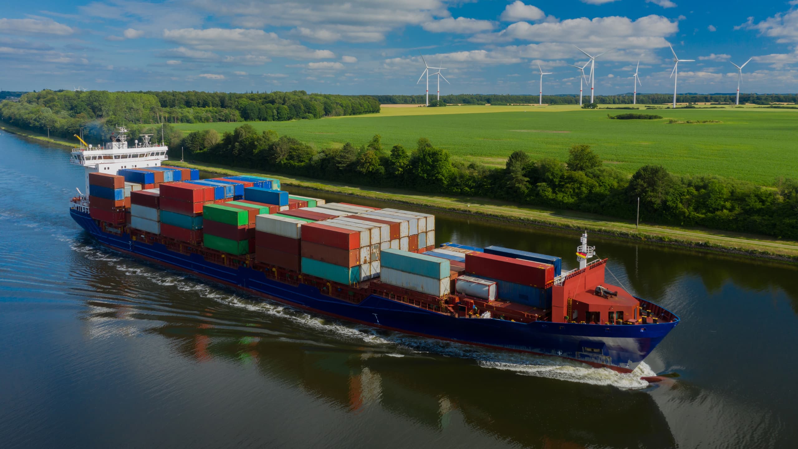 Cargo ship with multicolored containers in a waterway, with fields and wind turbines in the background.