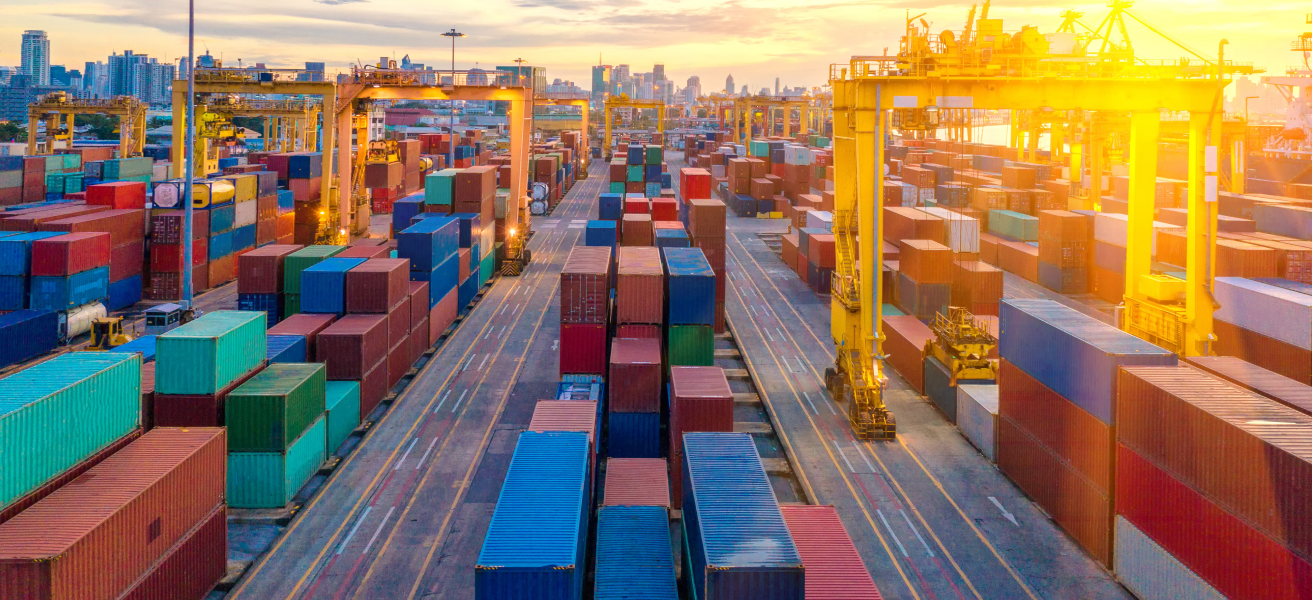 Busy shipping port with stacked containers and large yellow cranes at sunset.