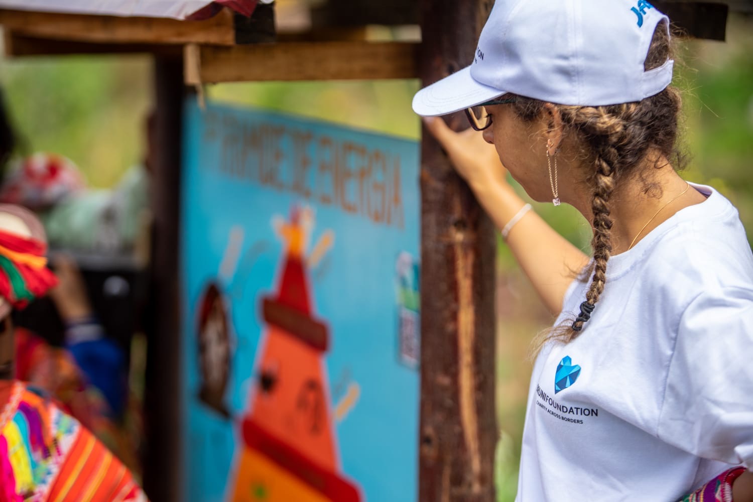 A volunteer wearing a white Bruni Foundation T-shirt and cap with braided hair observes a colorful educational display in an outdoor setting. The scene features vibrant colors and cultural elements, with blurred figures in traditional attire in the background.