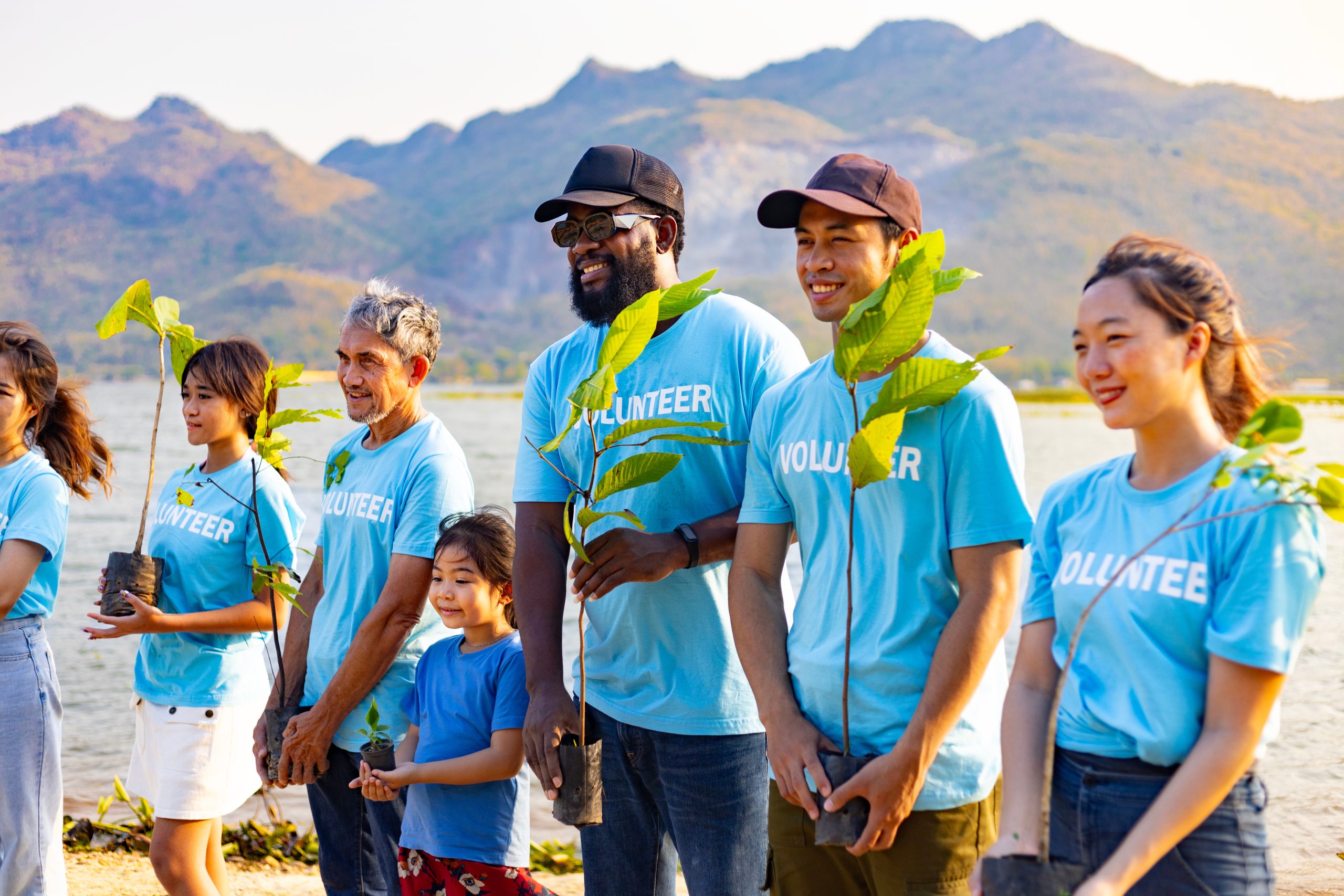 A diverse group of volunteers holding tree saplings near a lake with mountains in the background.
