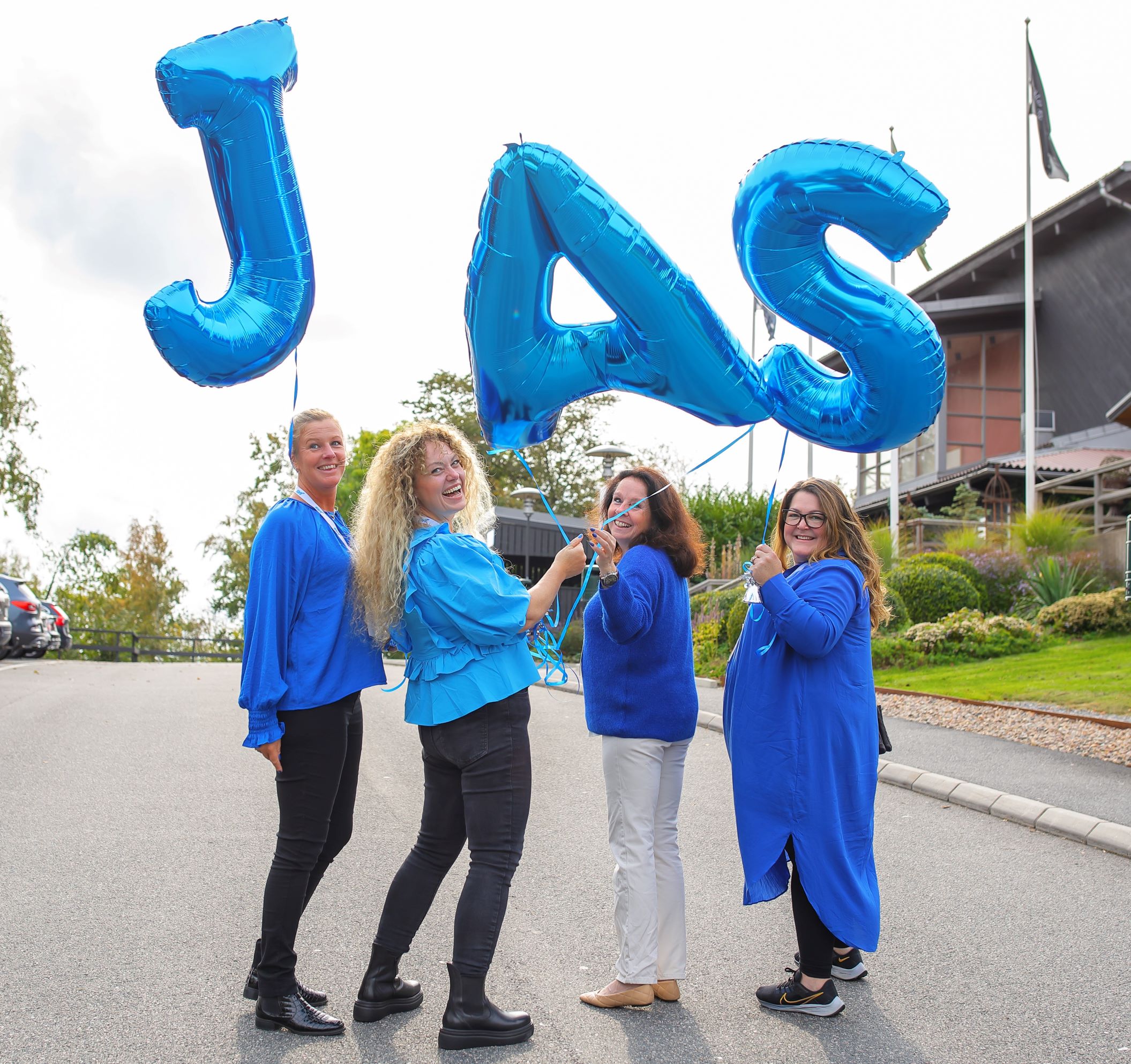 Four women standing in a line, looking back at the camera, holding blue mylar balloons that read, "JAS"