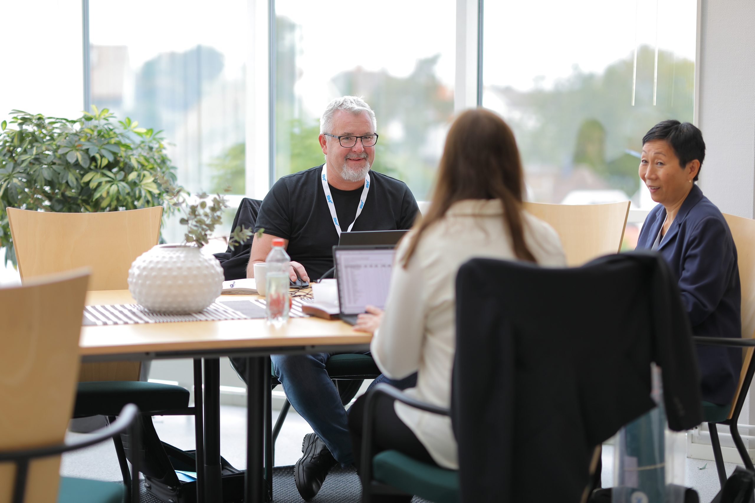 Three colleagues sitting around a table in a bright office with large windows, engaged in a discussion. A man with glasses and a lanyard smiles while using a laptop, facing two women. One woman is partially seen from behind, while the other, dressed in a blazer, smiles in conversation. The table features laptops, a decorative vase, and drinks.