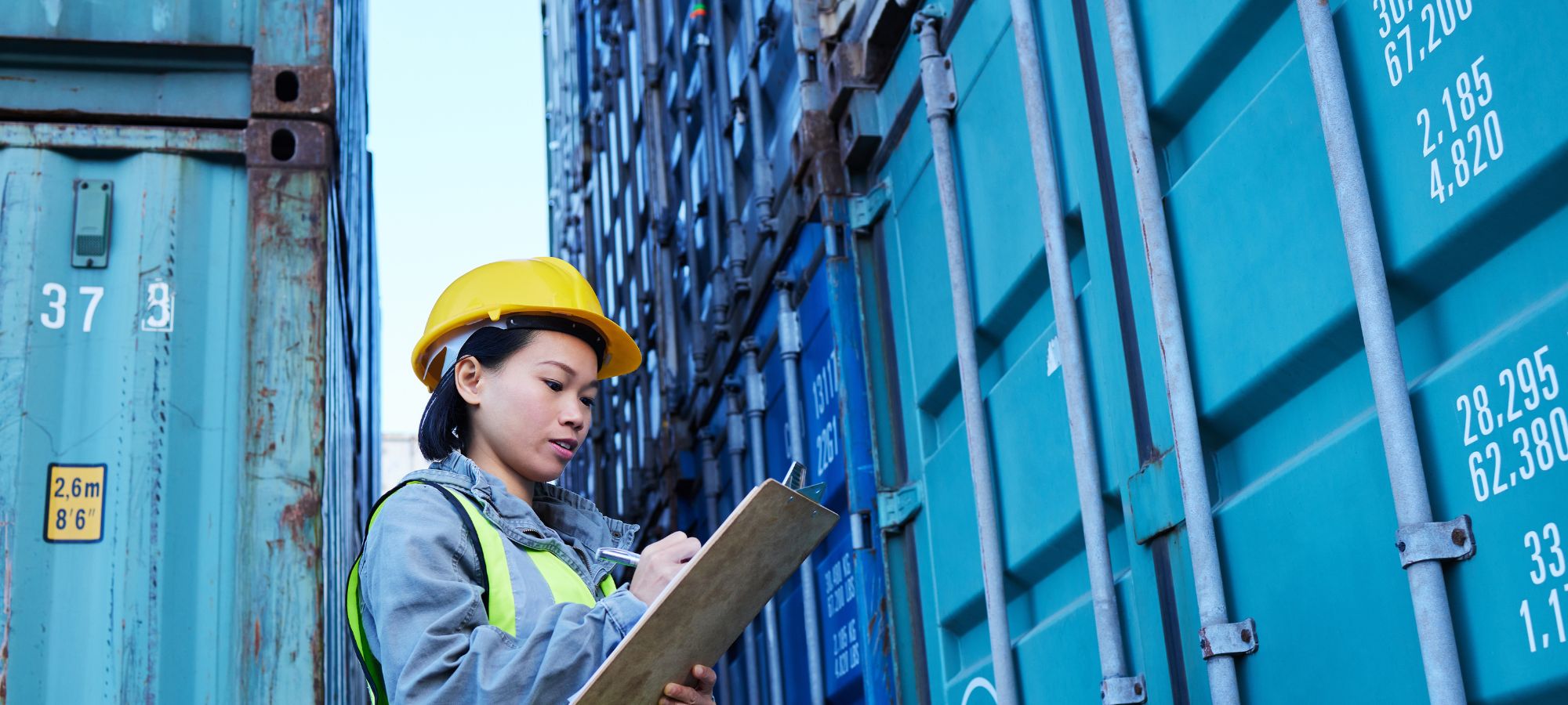 A logistics worker wearing a yellow hard hat and reflective vest checks a clipboard while inspecting large blue shipping containers stacked outdoors.