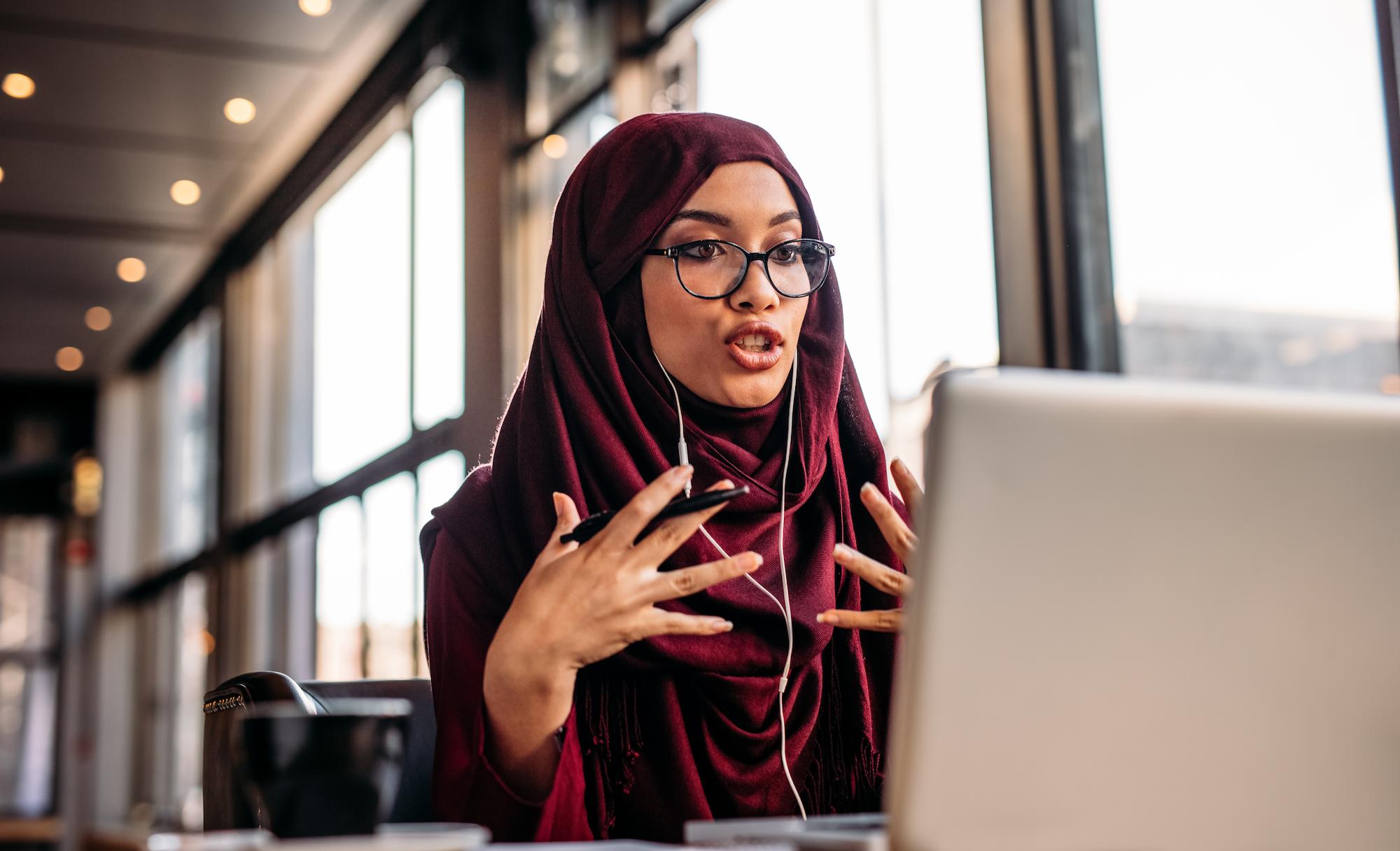 A woman in a hijab engages in conversation while using her laptop, showcasing a moment of communication and connection.