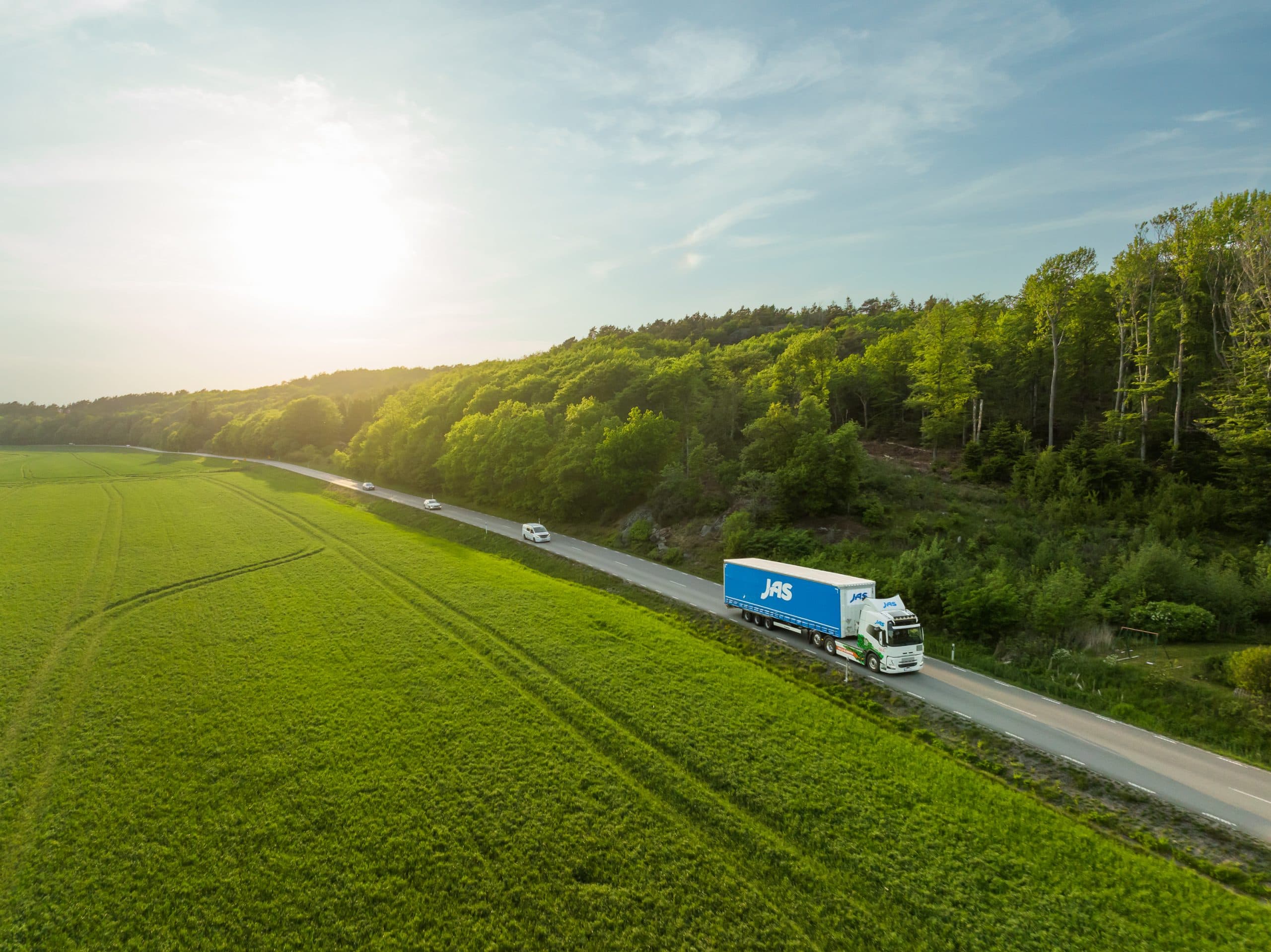 A truck with a blue "JAS" trailer driving on a road through green countryside under a sunny sky.
