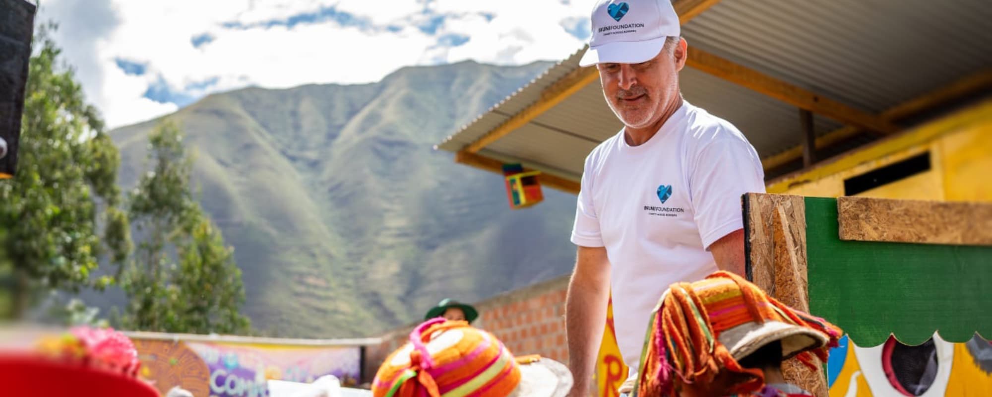 A person in a white shirt and cap interacts with others wearing colorful hats in a mountainous outdoor setting.