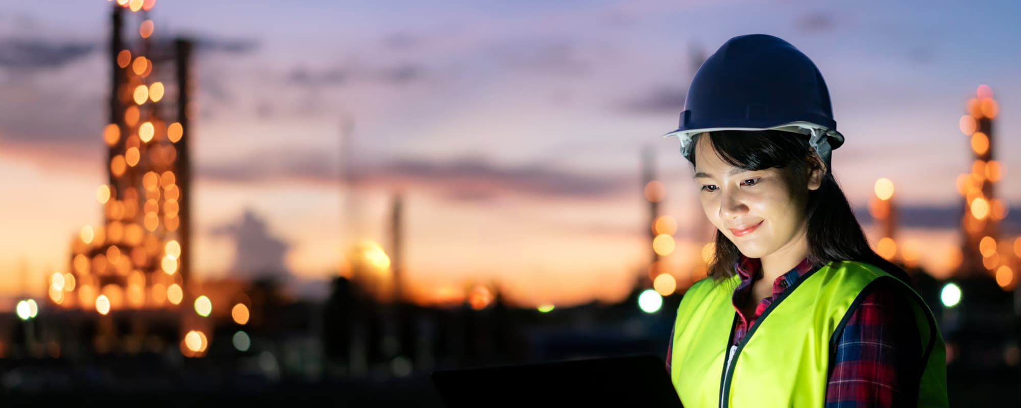 A woman in a safety helmet and vest smiles at a device in an industrial setting at dusk.