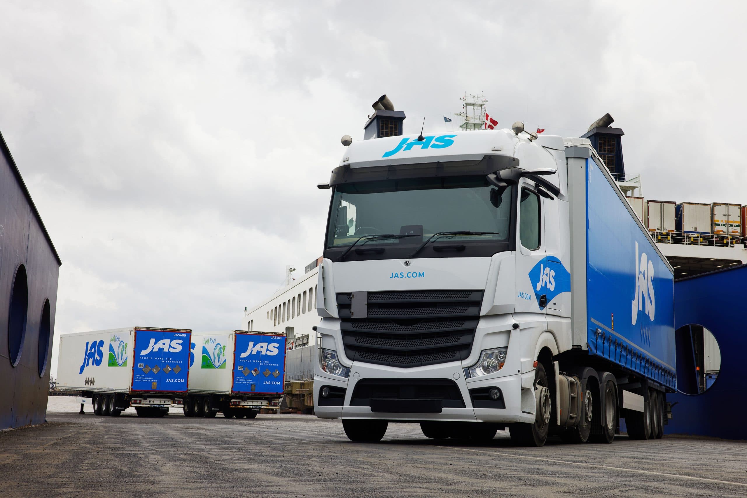 A freight truck with the "JAS" logo parked in front of a ship under a cloudy sky.