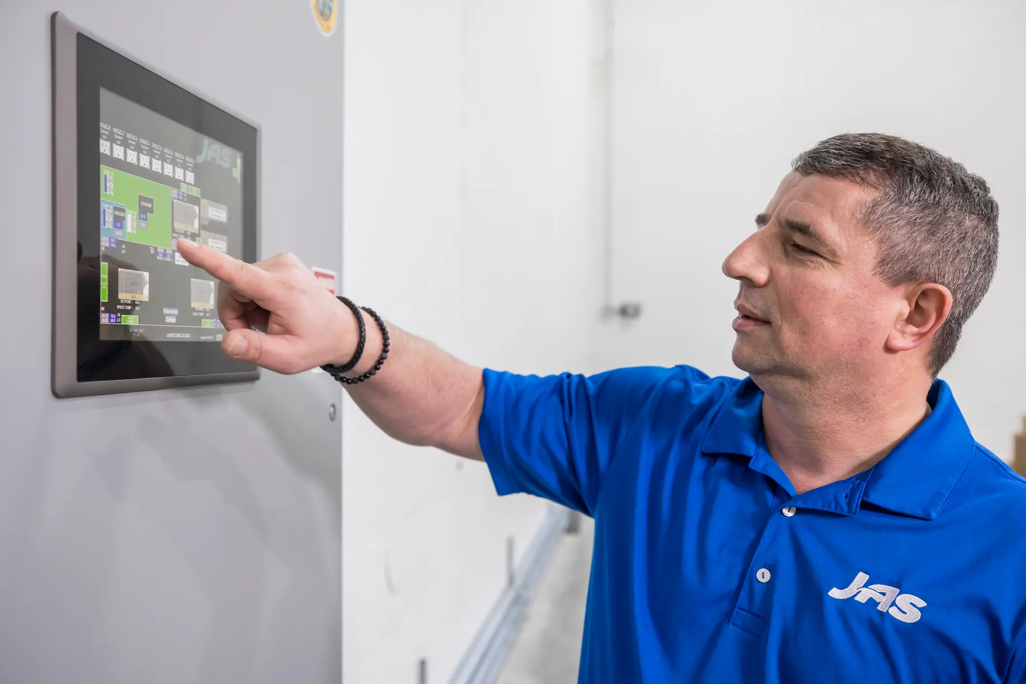 A man wearing a blue JAS-branded polo shirt interacts with a touchscreen control panel, showcasing logistics or operational technology in a professional environment.
