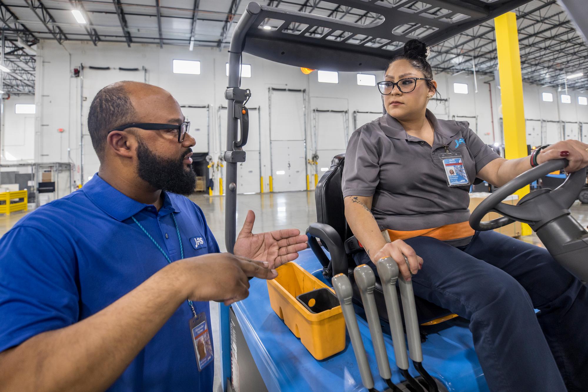 A man in a blue JAS-branded shirt gestures while speaking with a woman seated on industrial equipment inside a spacious warehouse environment.