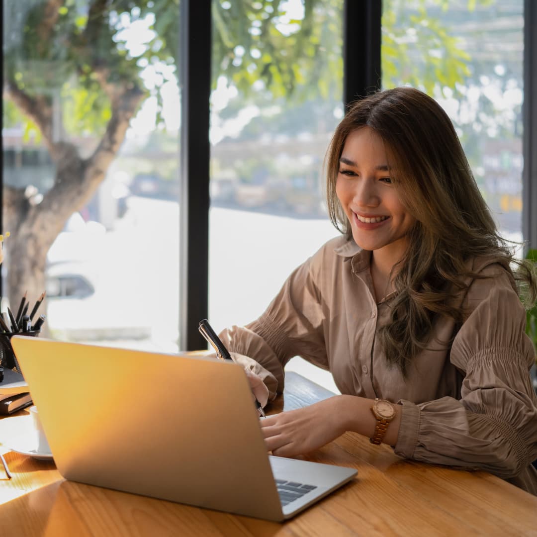 A woman smiling while working on a laptop and writing in a notebook at a wooden table, with large windows and a view of a tree outside.