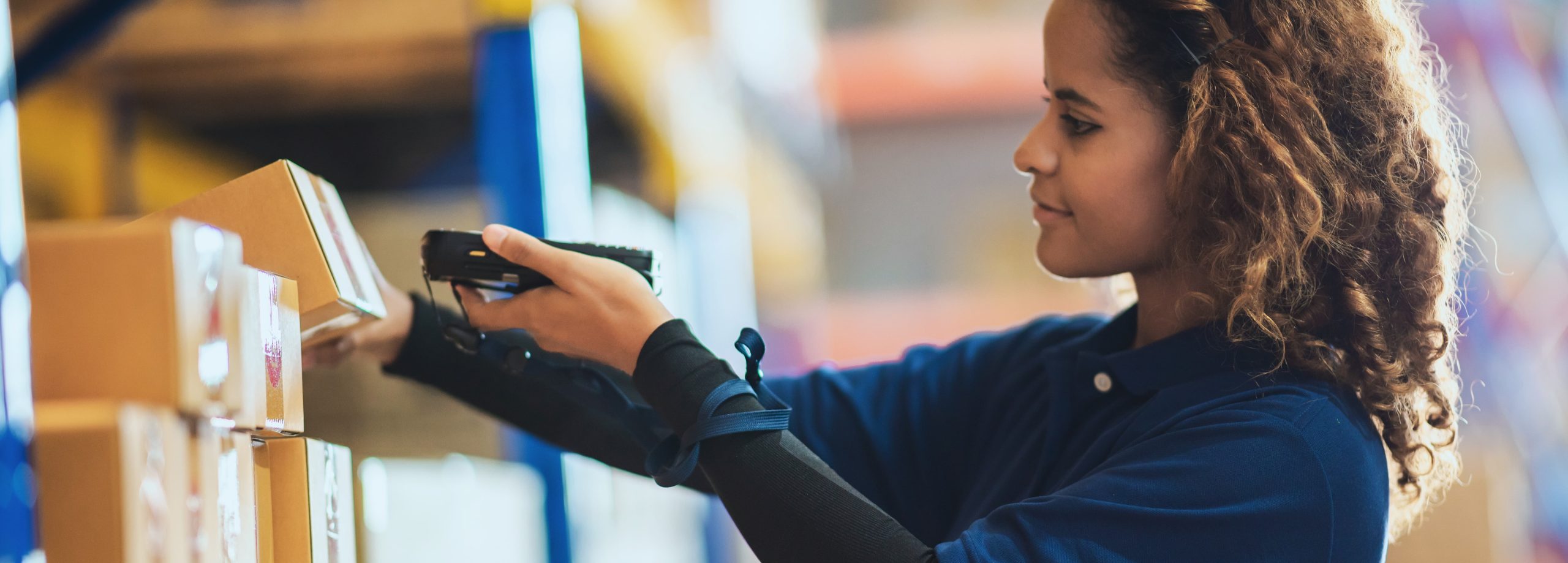 A woman using a handheld scanner in a warehouse, surrounded by stacked cardboard boxes.