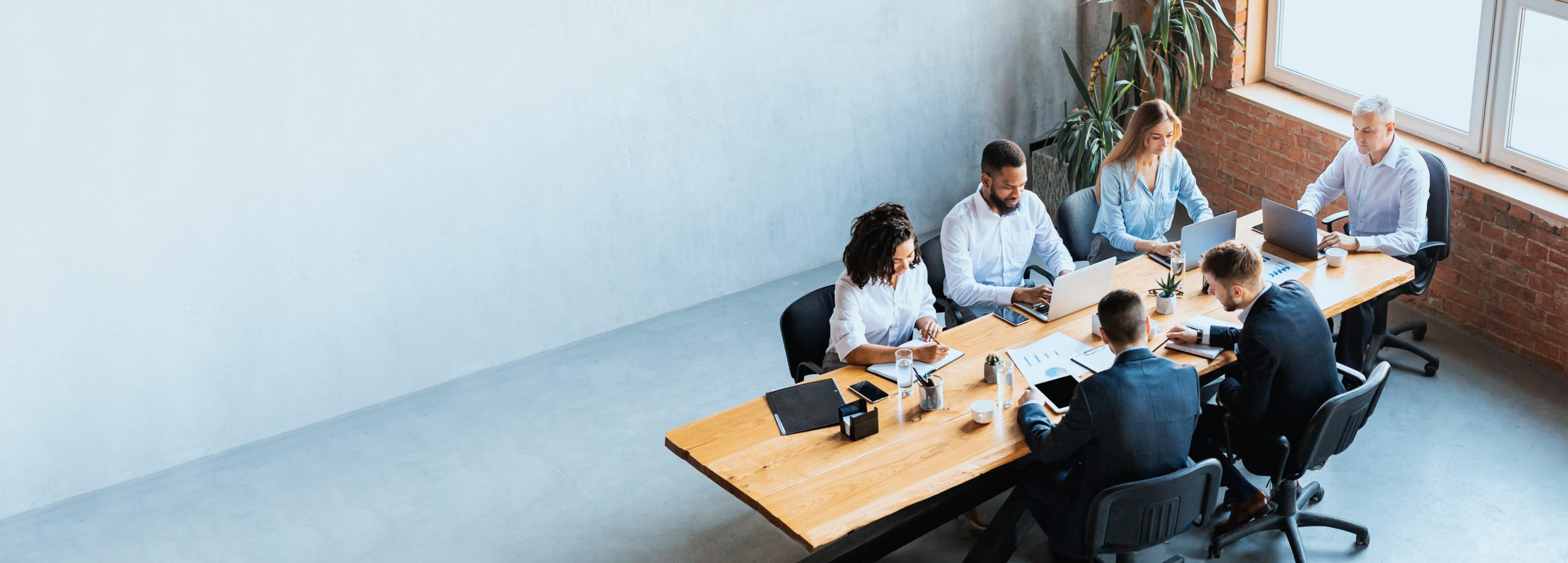 Office meeting with six people around a wooden table, using laptops and taking notes.