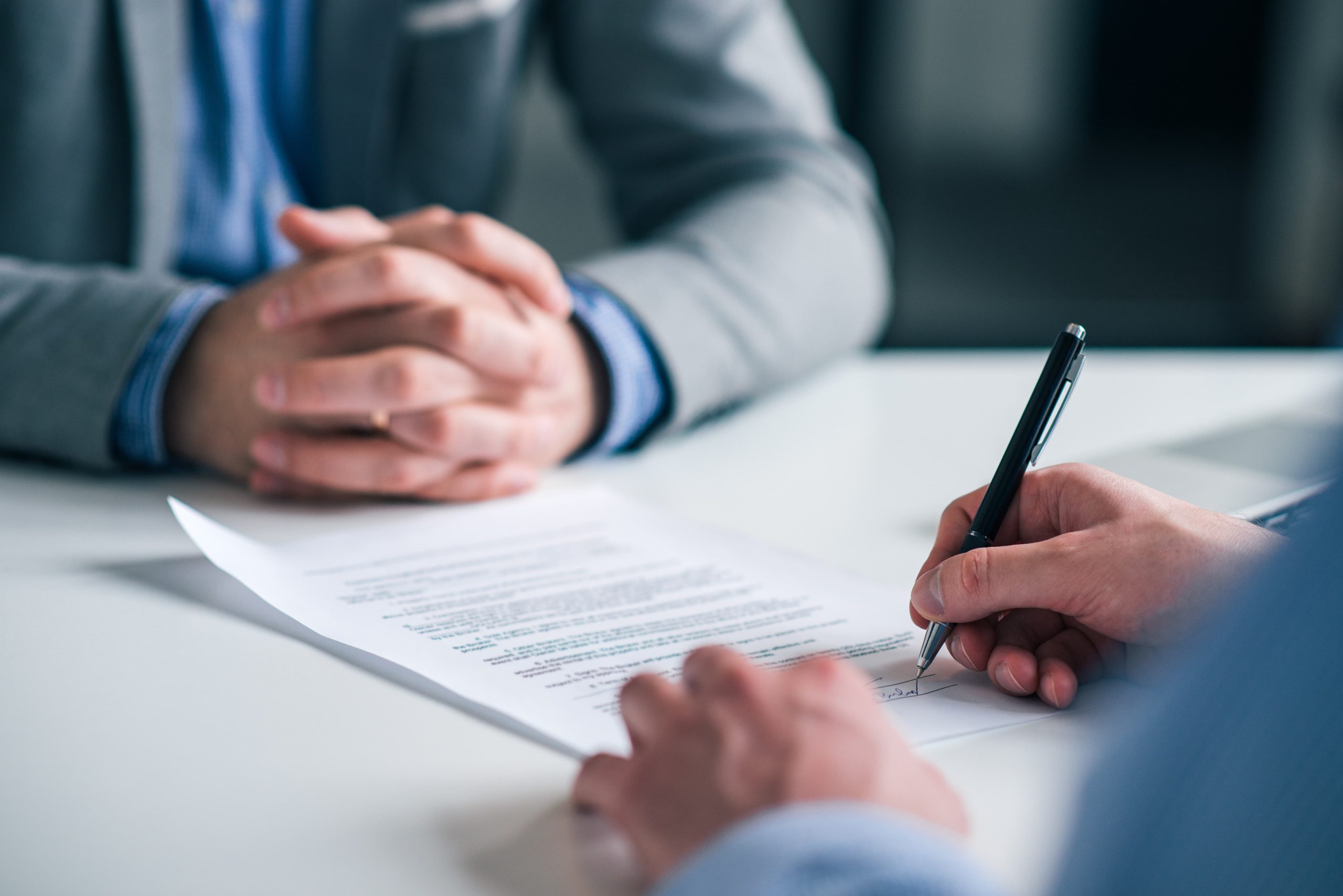 Close-up of a person signing a document in a professional setting, with another person's clasped hands visible nearby.