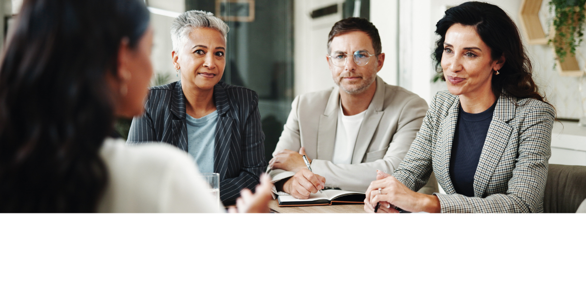 Four people sit at a meeting table, three facing the camera, engaged in discussion.